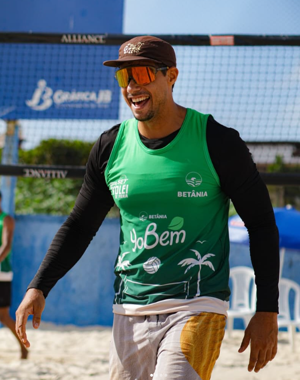 Bruno sorrindo, de camisa verde, boné e óculos alaranjados